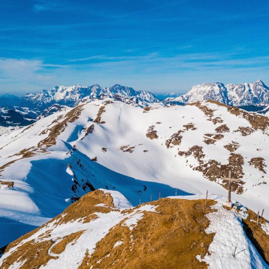 A mesmerizing shot of snow-covered mountains with a wooden cross on one of the peaks under a blue sky